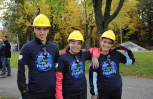 Three people wearing yellow hard hats and protective glasses stand outdoors. They are all smiling and wearing matching navy blue shirts that read "Travailler pour la planète." Trees with autumn foliage are in the background. One person holds a shovel, and another has a map.